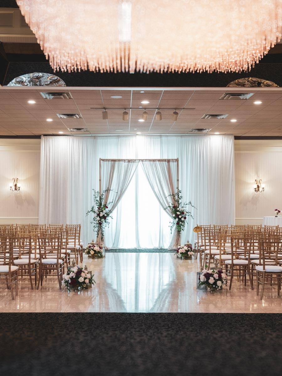 Elegant reception room at The Mirage with white drapings, curved aisle decorated with fresh greenery, signature chandelier, and rows of golden chiavari chairs