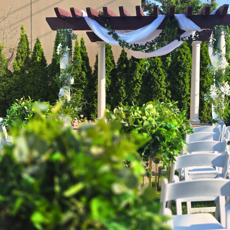 Wedding gazebo Michigan at The Mirage Clinton Township featuring a dark wood pergola with white draping and gold lantern lined aisle surrounded by arborvitae evergreens