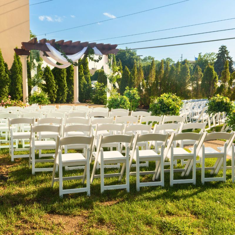 Outdoor ceremony seating at The Mirage wedding gazebo Michigan with white folding chairs facing a draped pergola stage surrounded by lush evergreen trees
